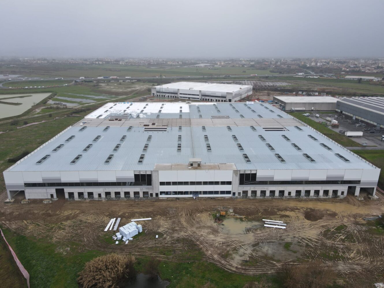 Un grande capannone industriale con un tetto bianco e un paesaggio incompiuto, un terreno fangoso, materiali da costruzione ed edifici vicini.