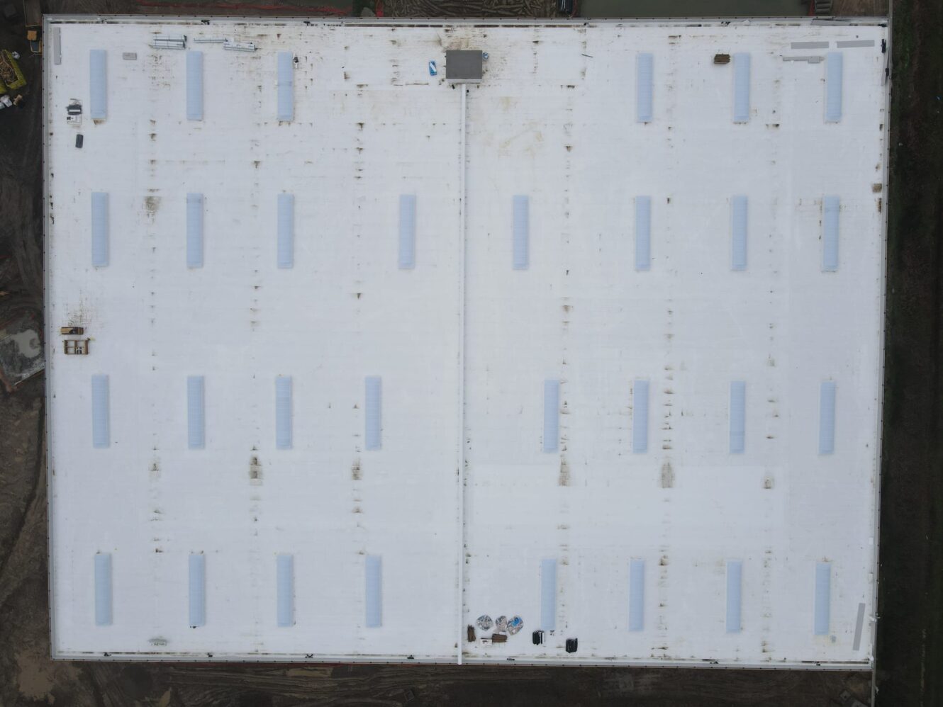 Vista aerea del tetto di un grande edificio industriale bianco e piatto, caratterizzato da lucernari rettangolari uniformemente distanziati e da leggeri segni di sporco.