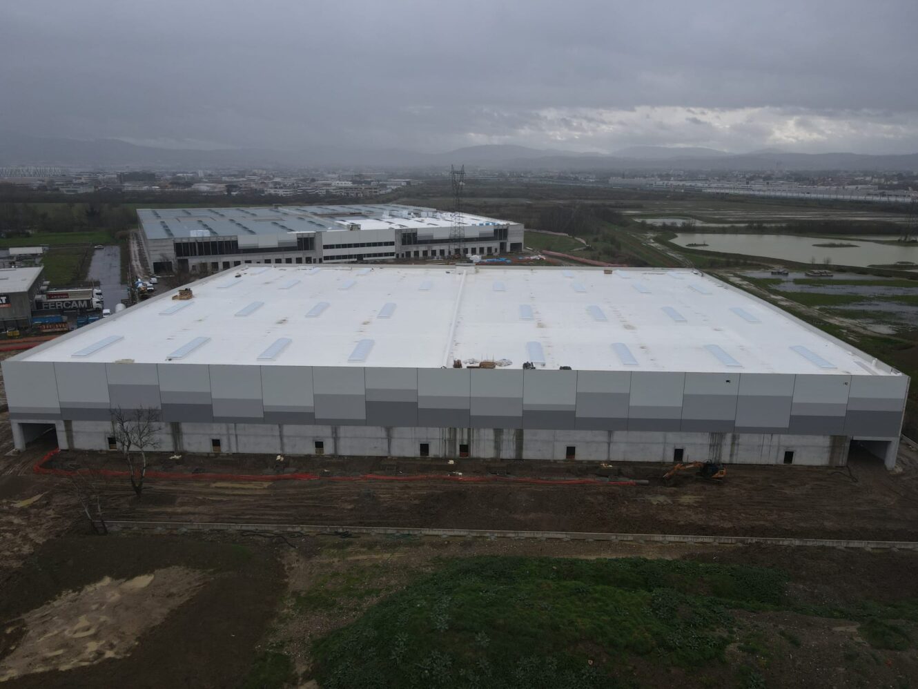 Un grande magazzino rettangolare con tetto bianco e pareti a strisce grigie, terreno umido intorno, accanto ad altri edifici industriali.