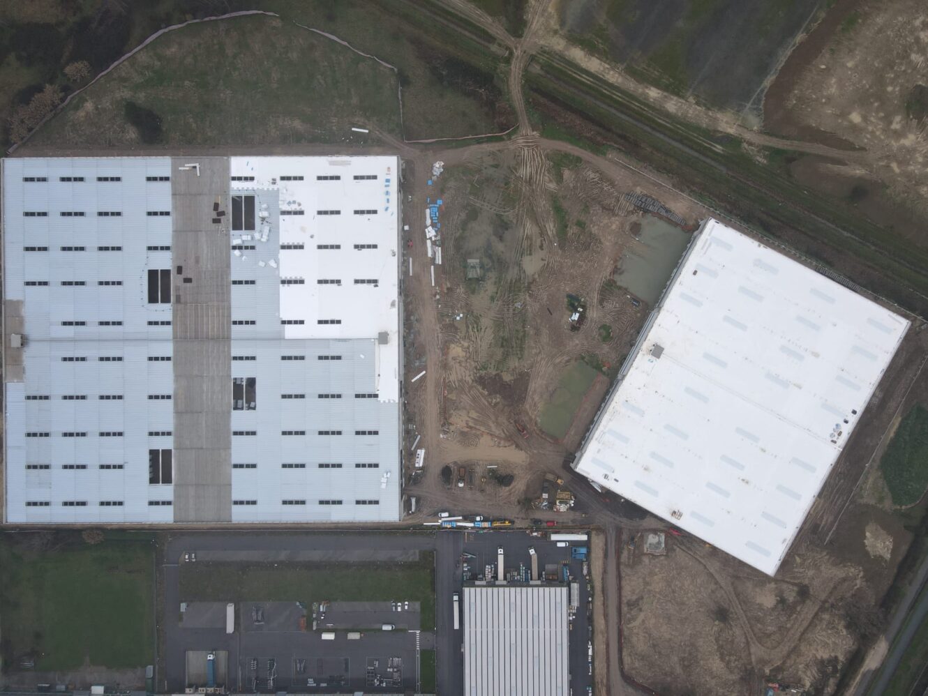 Una veduta aerea mostra due grandi edifici industriali separati da un cantiere con attrezzature e percorsi sterrati visibili.