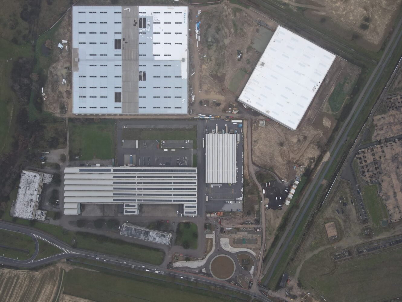 Veduta aerea di un complesso industriale con grandi capannoni rettangolari, aree di parcheggio, strade circostanti e terreni aperti nelle vicinanze.