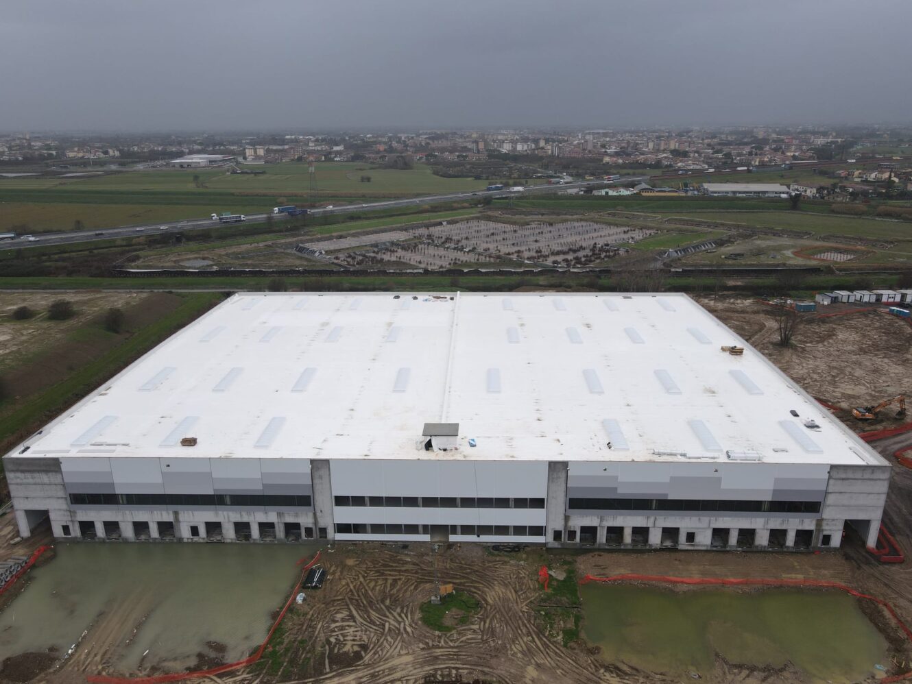 Un grande magazzino rettangolare con il tetto bianco è in costruzione, circondato da terreno fangoso, pozzanghere, campi e una città lontana.