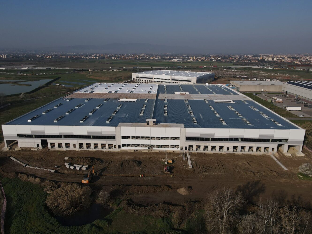 Veduta aerea di un grande magazzino industriale in costruzione, con veicoli e attrezzature da cantiere davanti.