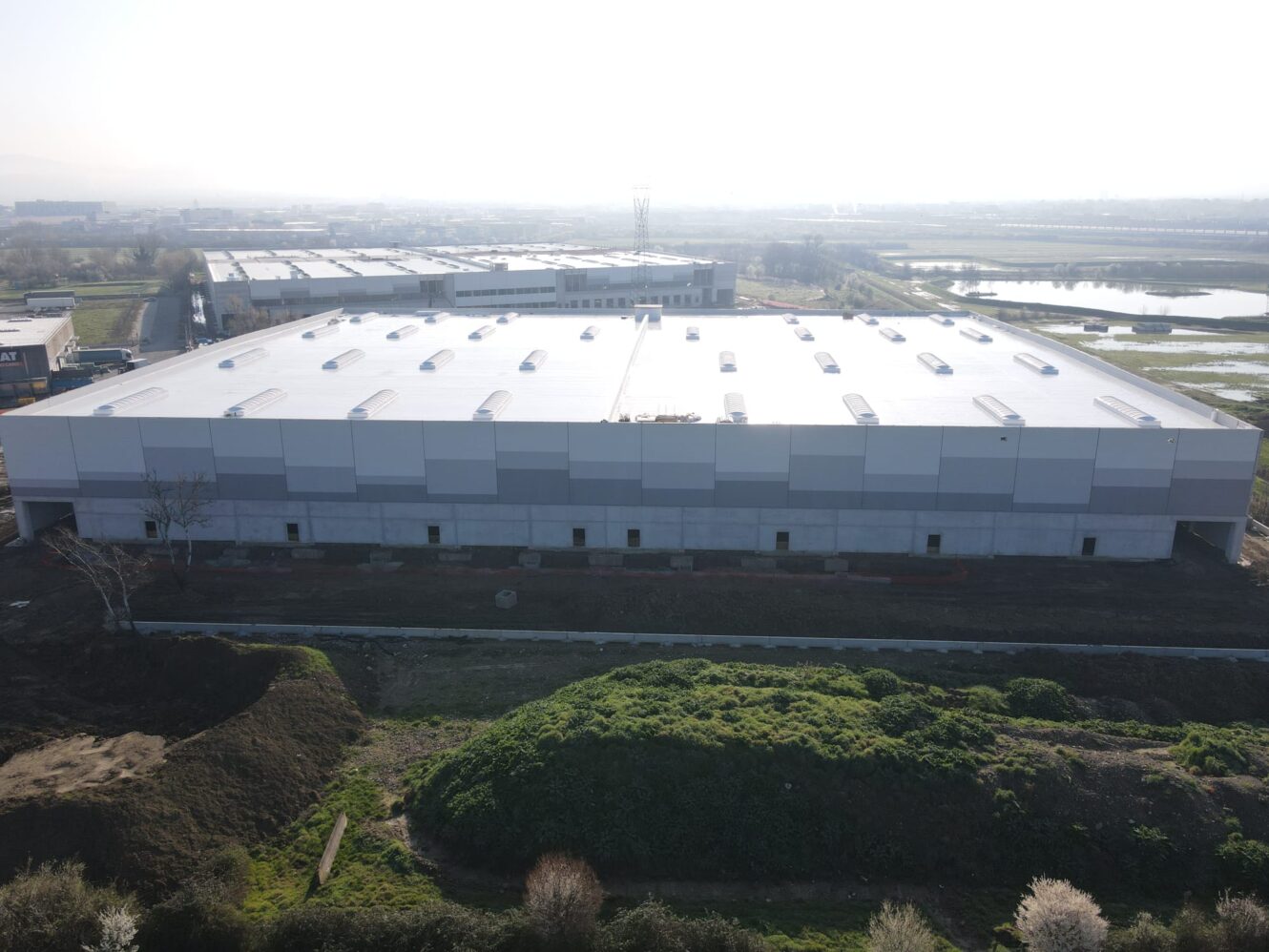 Un grande magazzino industriale rettangolare con tetto piatto e lucernari multipli, aree erbose ed edifici vicini sotto un cielo nebuloso.