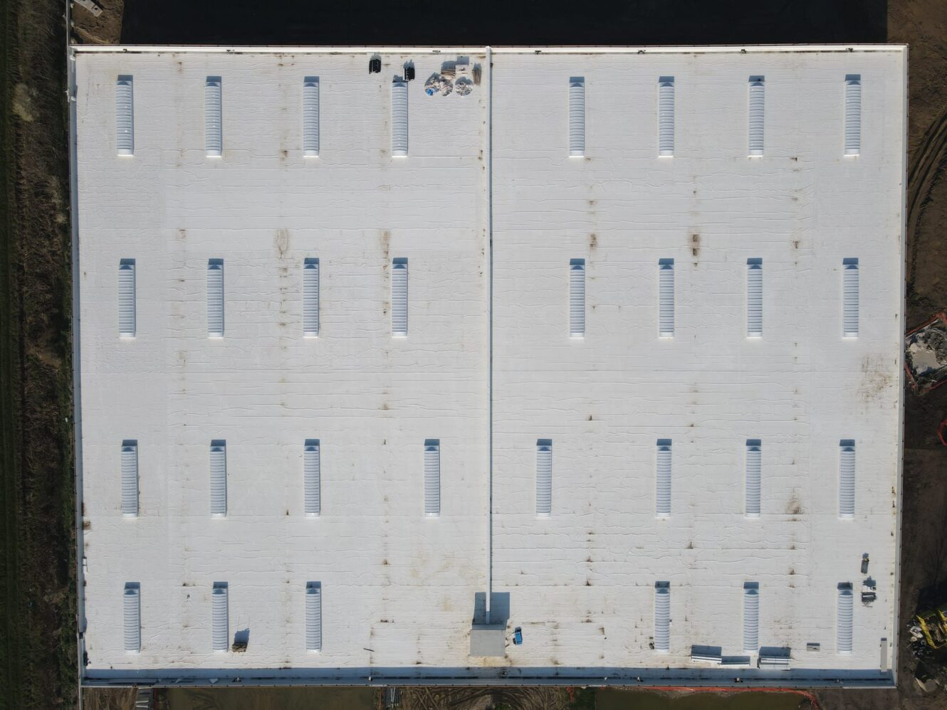 Vista aerea del tetto di un edificio industriale bianco con lucernari rettangolari uniformemente distanziati e attrezzature raggruppate vicino al centro.