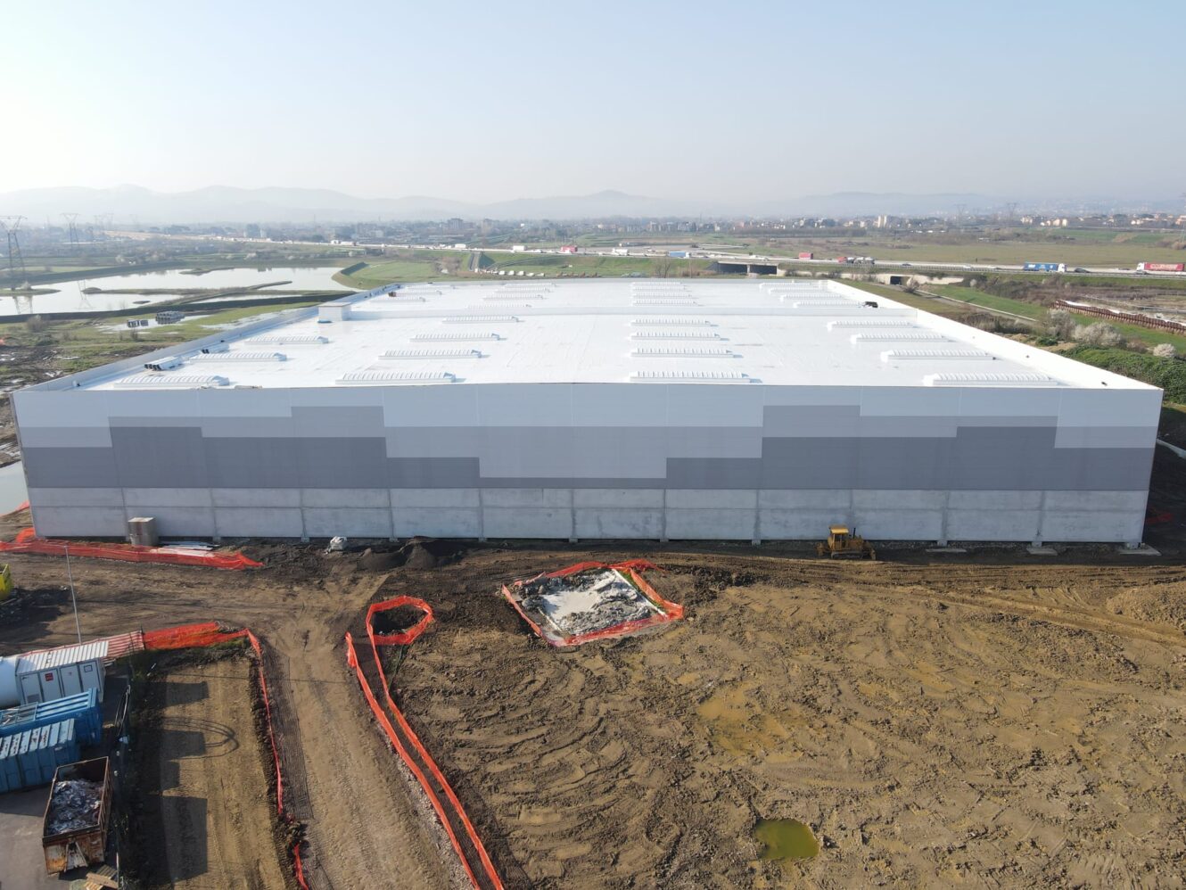 Un grande magazzino industriale rettangolare in costruzione, circondato da terra, macchinari, barriere di sicurezza, campi e una città alle spalle.