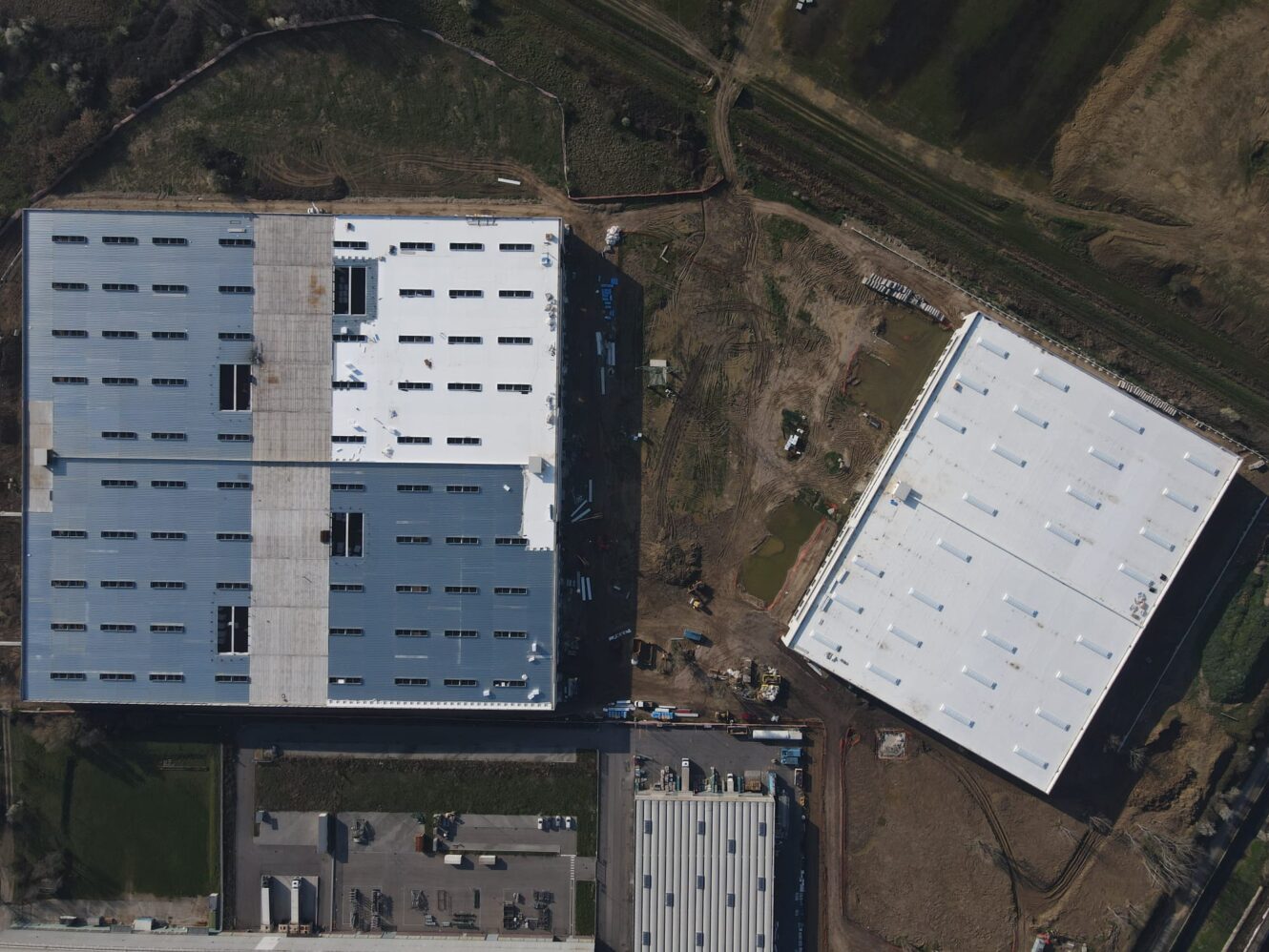 Vista aerea di grandi capannoni industriali con tetti grigi, sporcizia, erba, strade e attrezzature edili vicino all'edificio di destra.