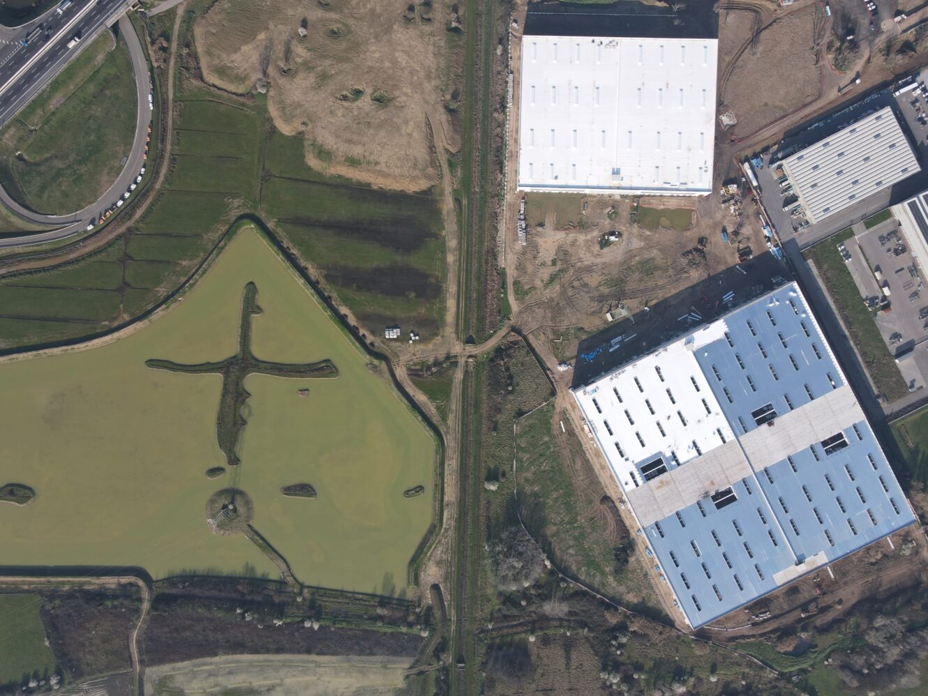 Vista aerea di due grandi edifici industriali bianchi accanto a uno stagno con piccole isole, circondato da strade e aree erbose.