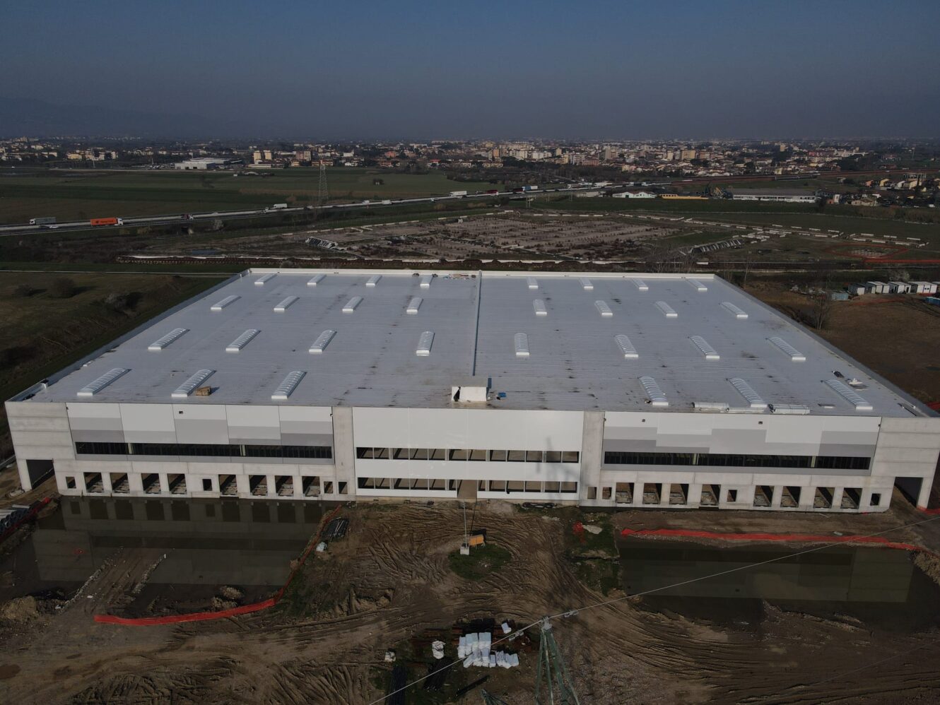 Un grande capannone industriale rettangolare in costruzione, circondato da terra e attrezzature, con un paesaggio urbano e colline alle spalle.
