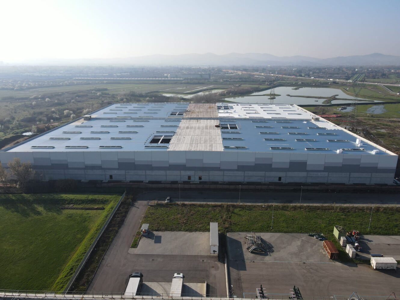 Un grande capannone industriale rettangolare con il tetto parzialmente aperto, circondato da campi, strade e canali d'acqua sotto un cielo nebuloso.