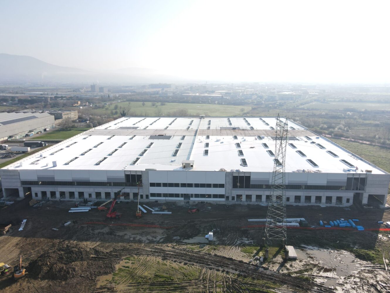 Veduta aerea di un grande capannone industriale dal tetto bianco in costruzione, con attrezzature e materiali in primo piano.