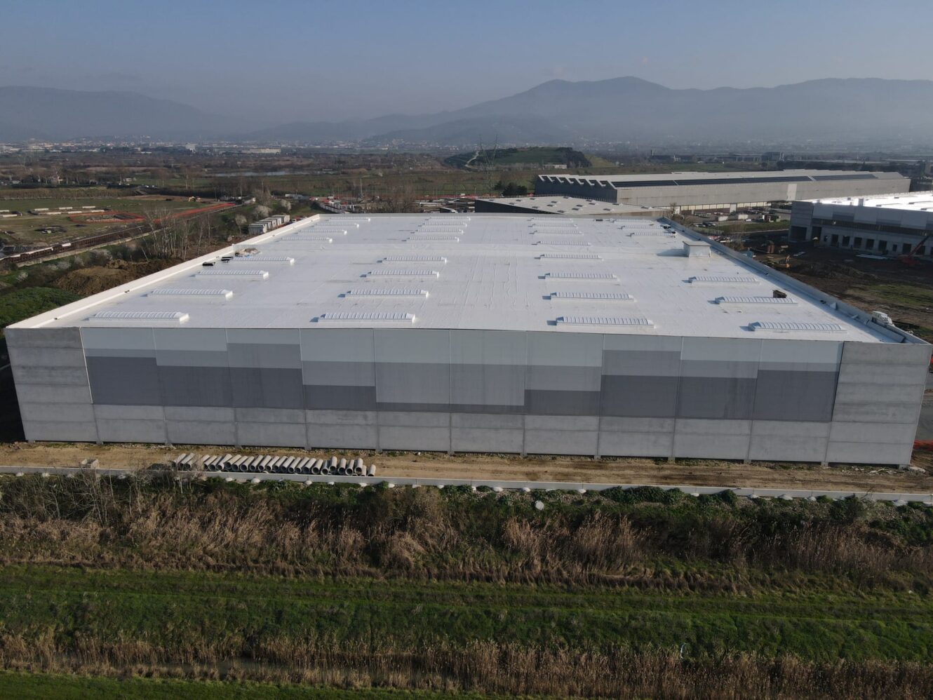 Un grande magazzino industriale rettangolare con tetto bianco e pareti grigie, circondato da campi ed erba, con montagne sullo sfondo.