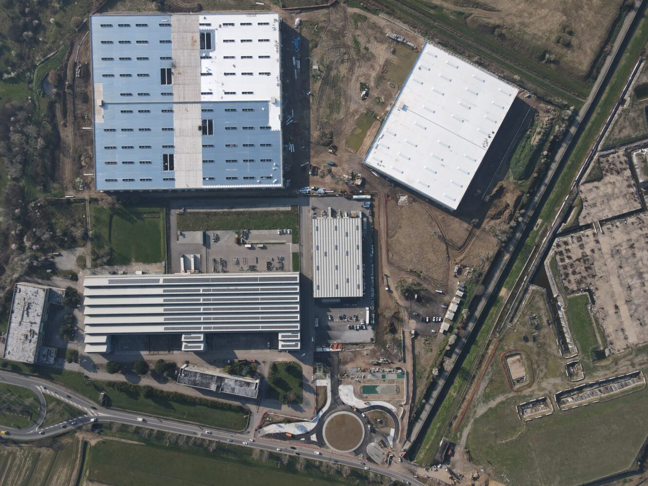 Vista aerea di edifici industriali con grandi tetti bianchi, strade circostanti, spazi verdi e cantieri vicini.