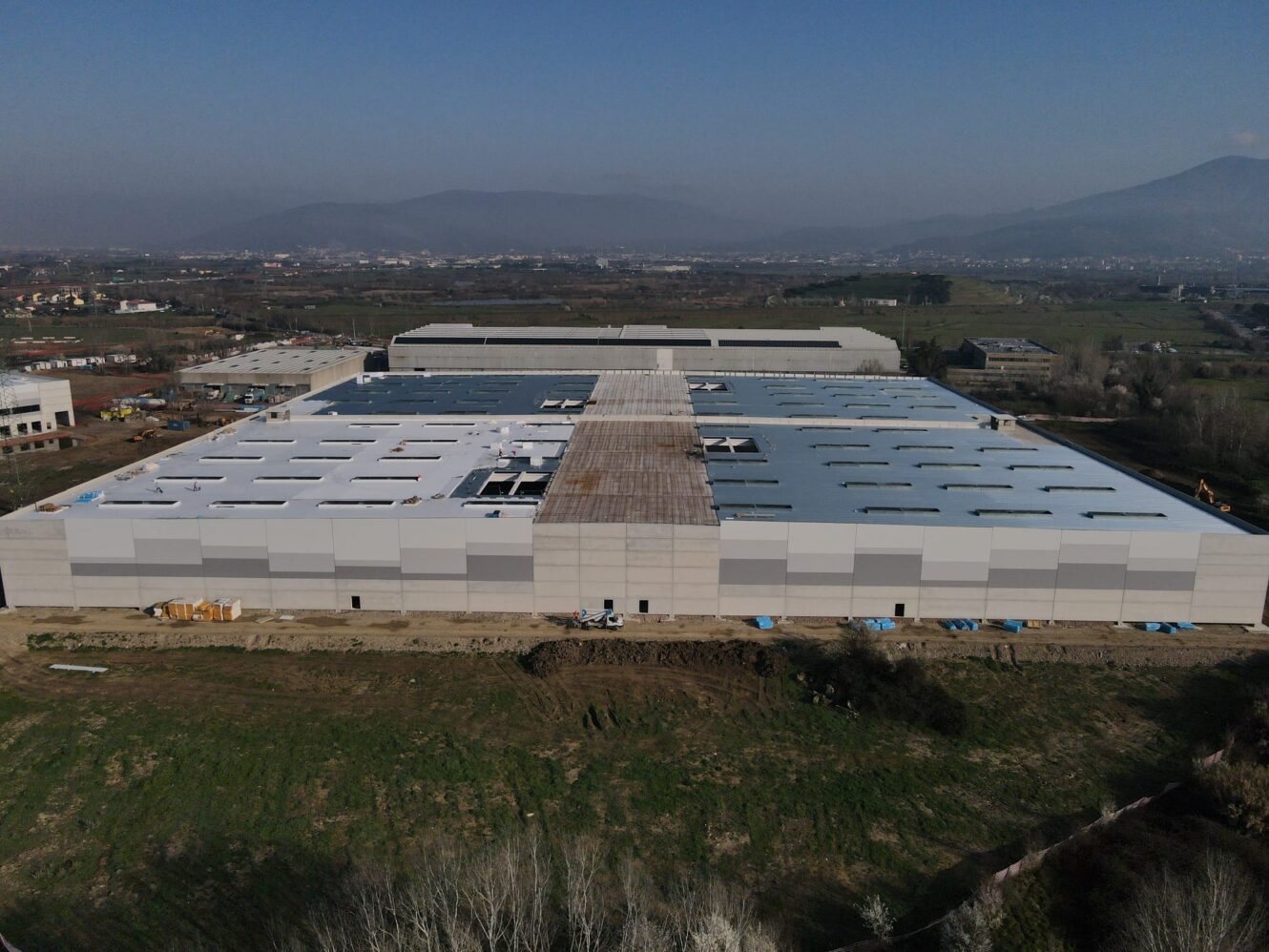 Veduta aerea di un grande edificio industriale rettangolare con tetto parzialmente completato, campi e montagne sotto un cielo sereno.