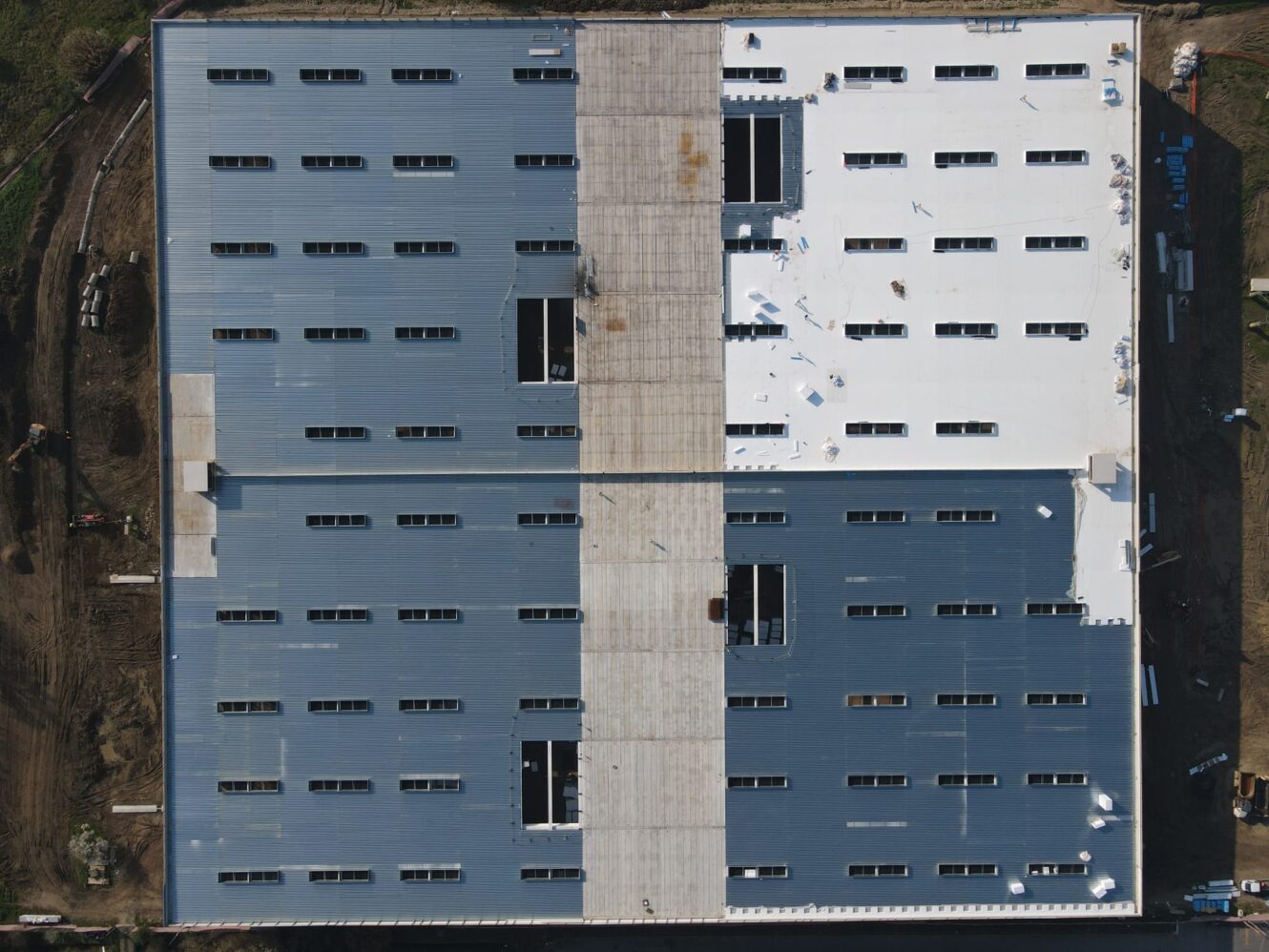 Vista aerea di un grande edificio rettangolare in costruzione, con tetto grigio, bianco e in cemento. Le attrezzature da costruzione lo circondano.
