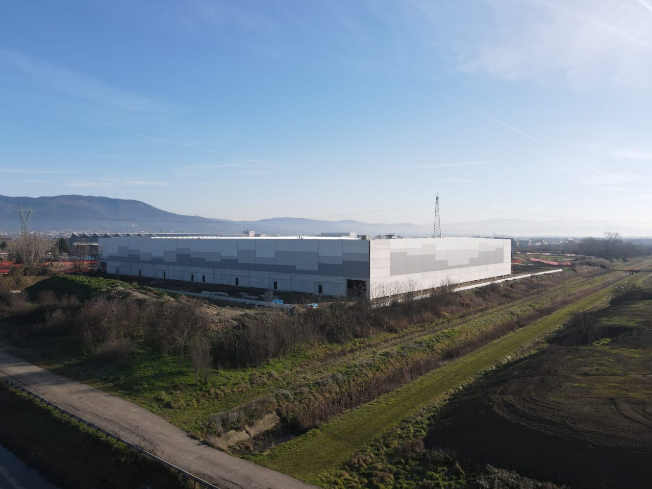 Un grande edificio industriale rettangolare con l'esterno grigio e bianco si trova in un'area rurale erbosa sotto un cielo limpido.