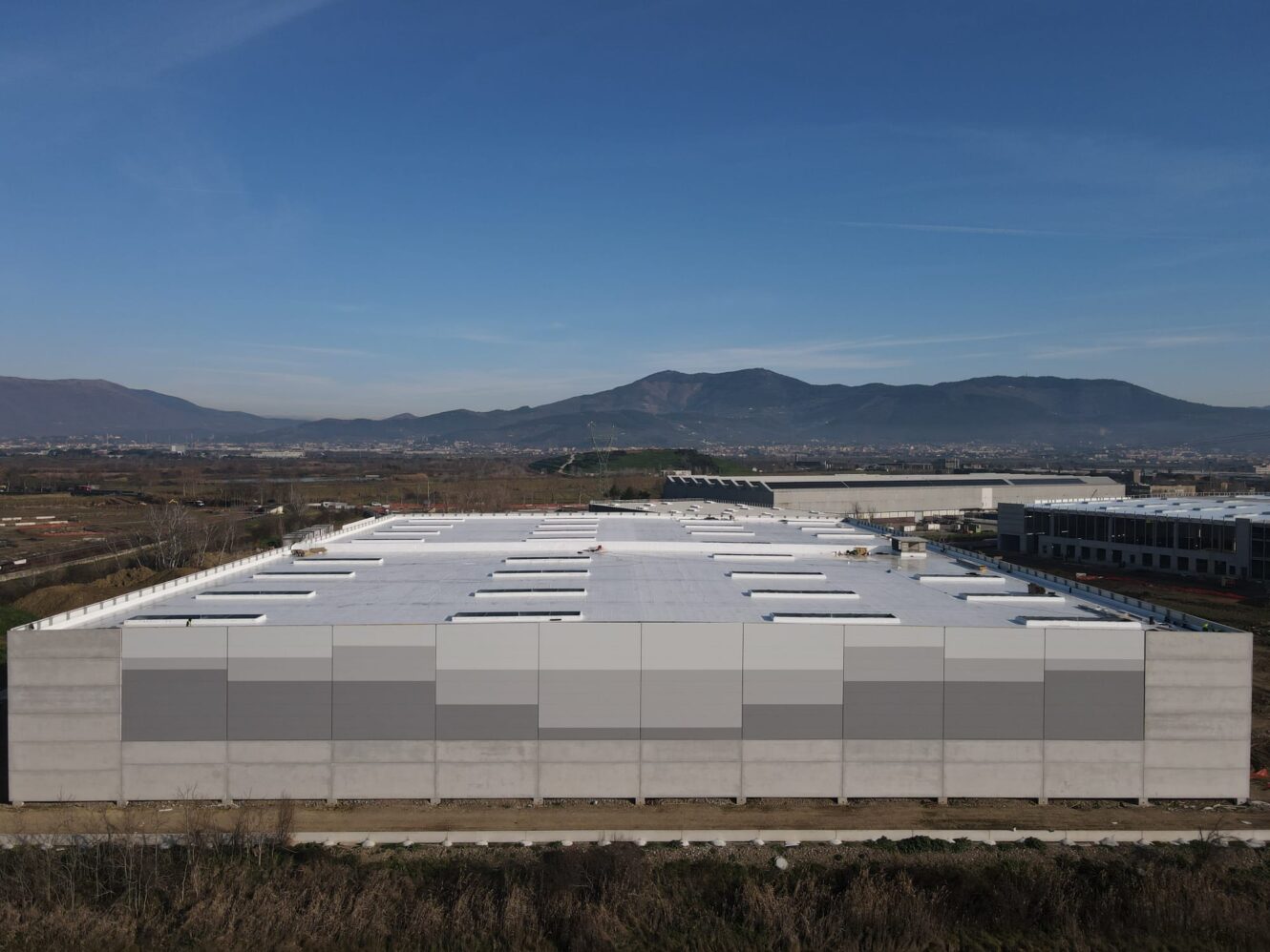 Un grande capannone industriale dal tetto bianco, circondato da campi, con montagne e un cielo azzurro sullo sfondo.