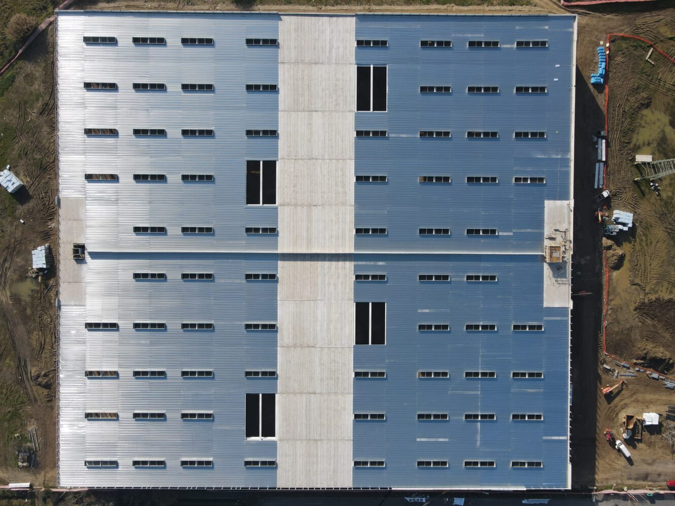 Veduta aerea di un grande edificio industriale rettangolare con tetto metallico e file di lucernari, circondato da attrezzature edili e terra.