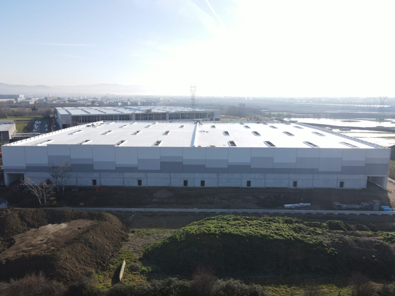 Un grande magazzino rettangolare con tetto piatto si trova su un terreno sgombro, circondato da materiali da costruzione e verde sotto un cielo limpido.