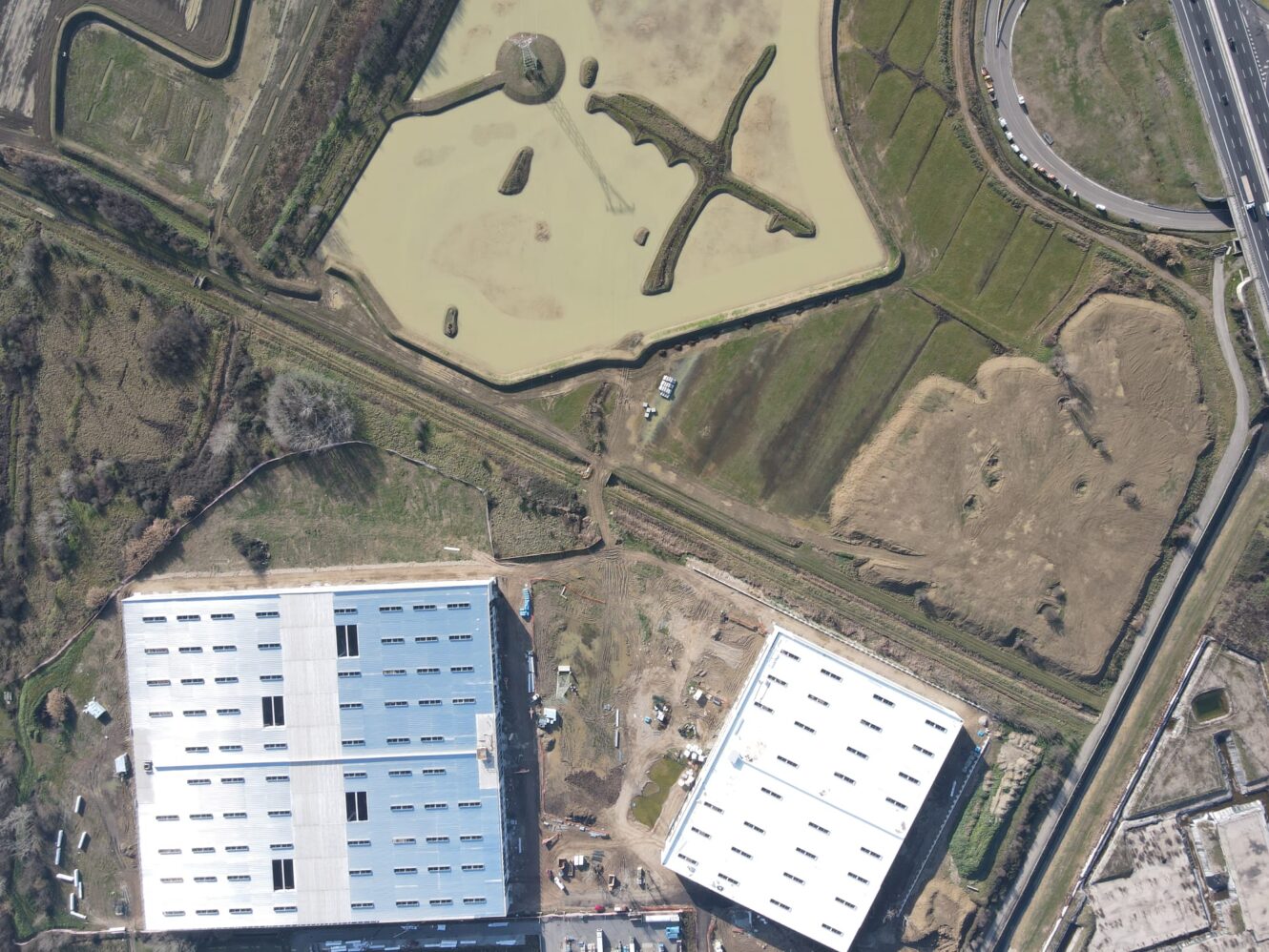 Veduta aerea di due grandi edifici industriali, attività di costruzione adiacenti, uno stagno con isole e strade e campi circostanti.