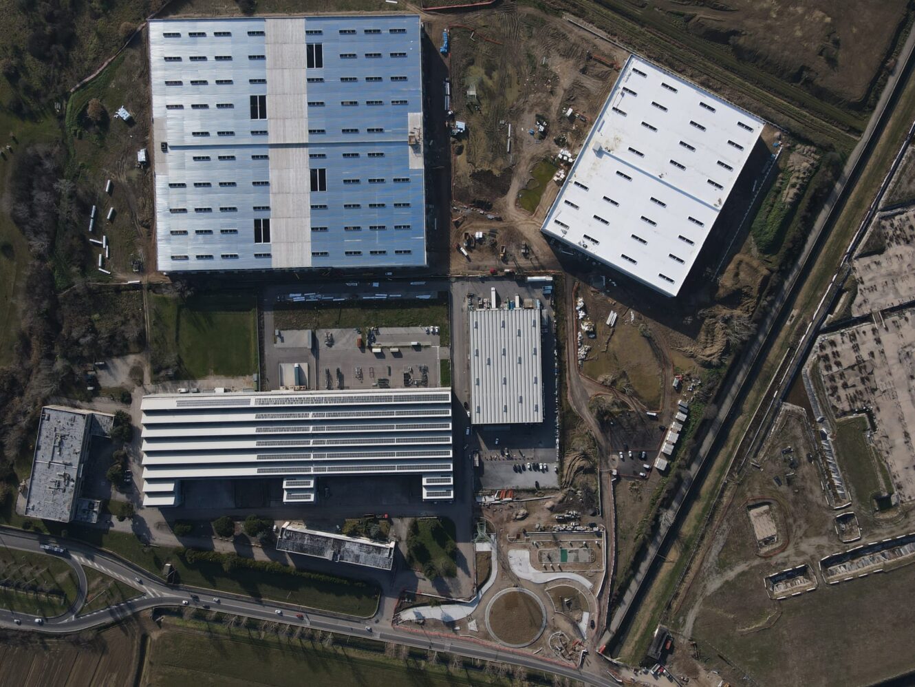 Veduta aerea di un complesso industriale con quattro grandi magazzini, strade adiacenti, aree di costruzione e spazi verdi circostanti.
