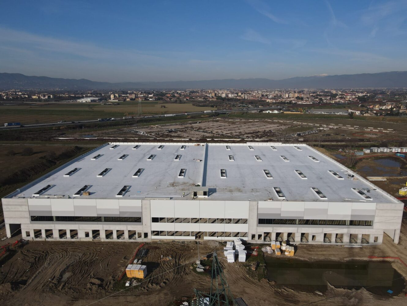 Un grande edificio industriale rettangolare con il tetto bianco è circondato da aree di costruzione sterrate, campi e un paesaggio urbano lontano, sotto un cielo azzurro e limpido.