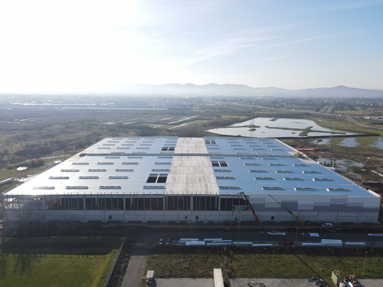 Un grande edificio industriale con tetto bianco in costruzione, circondato da campi, acqua e colline lontane sotto un cielo limpido.
