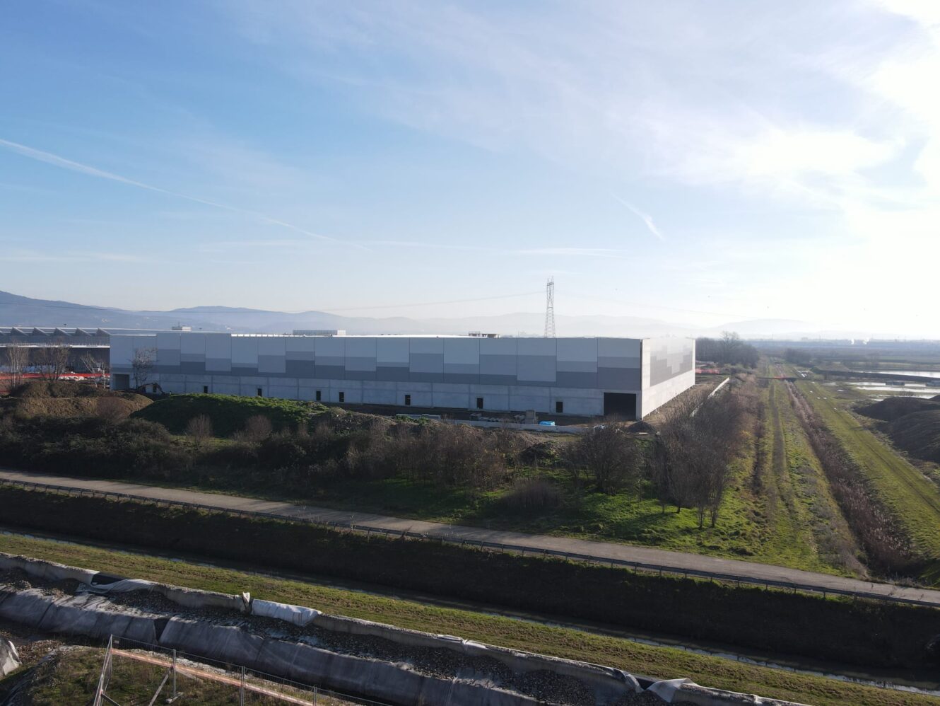 Un grande e moderno edificio adibito a magazzino con il tetto piatto si trova accanto a una strada e a zone erbose sotto un cielo limpido.