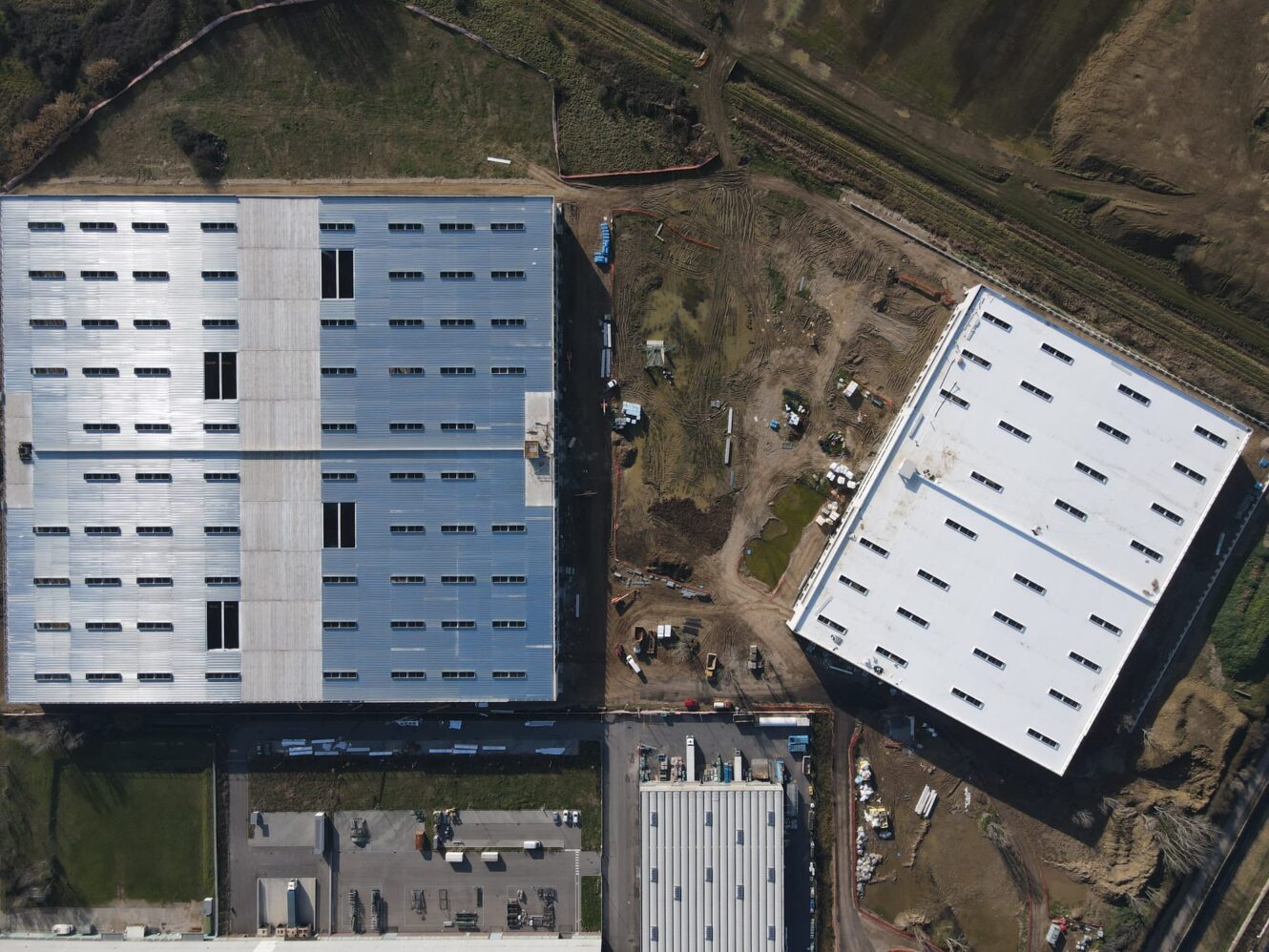Veduta aerea di due grandi edifici adibiti a magazzino, uno dei quali in costruzione, circondati da terra, macchinari e strade adiacenti.