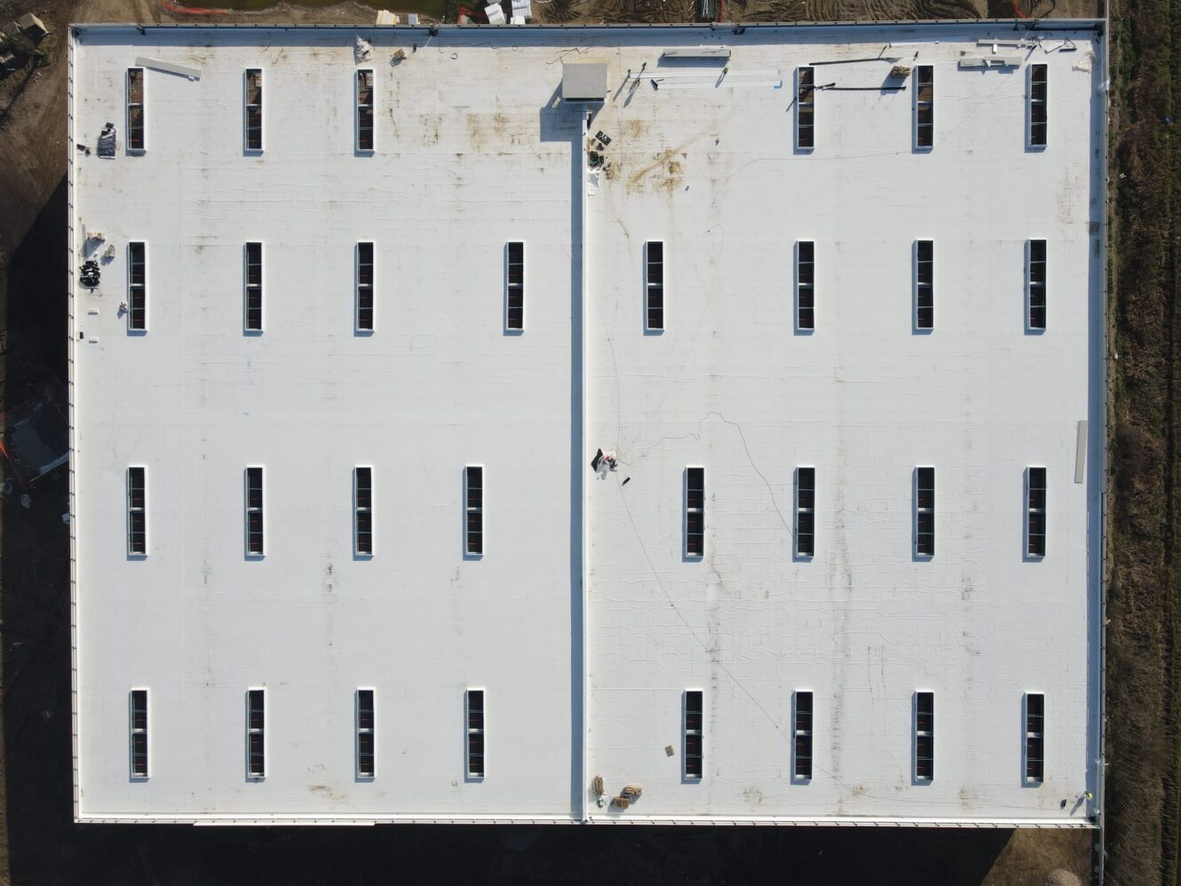 Vista aerea del tetto di un grande edificio rettangolare bianco con molteplici lucernari rettangolari uniformemente distanziati e addetti alla manutenzione visibili.