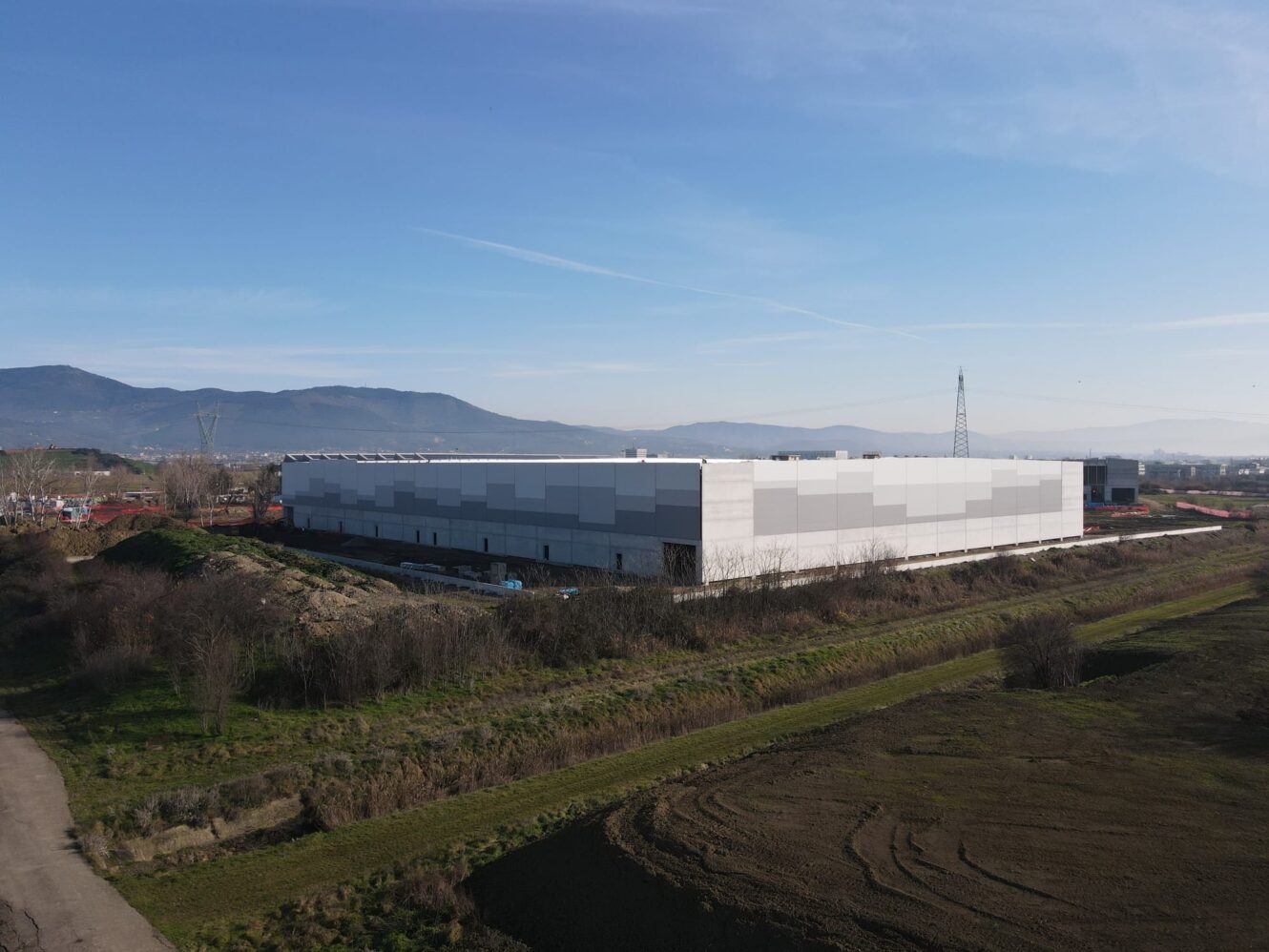 Un grande e moderno edificio industriale con tetto piatto situato in una zona semi-rurale, circondato da erba e vegetazione rada, con le montagne sullo sfondo.