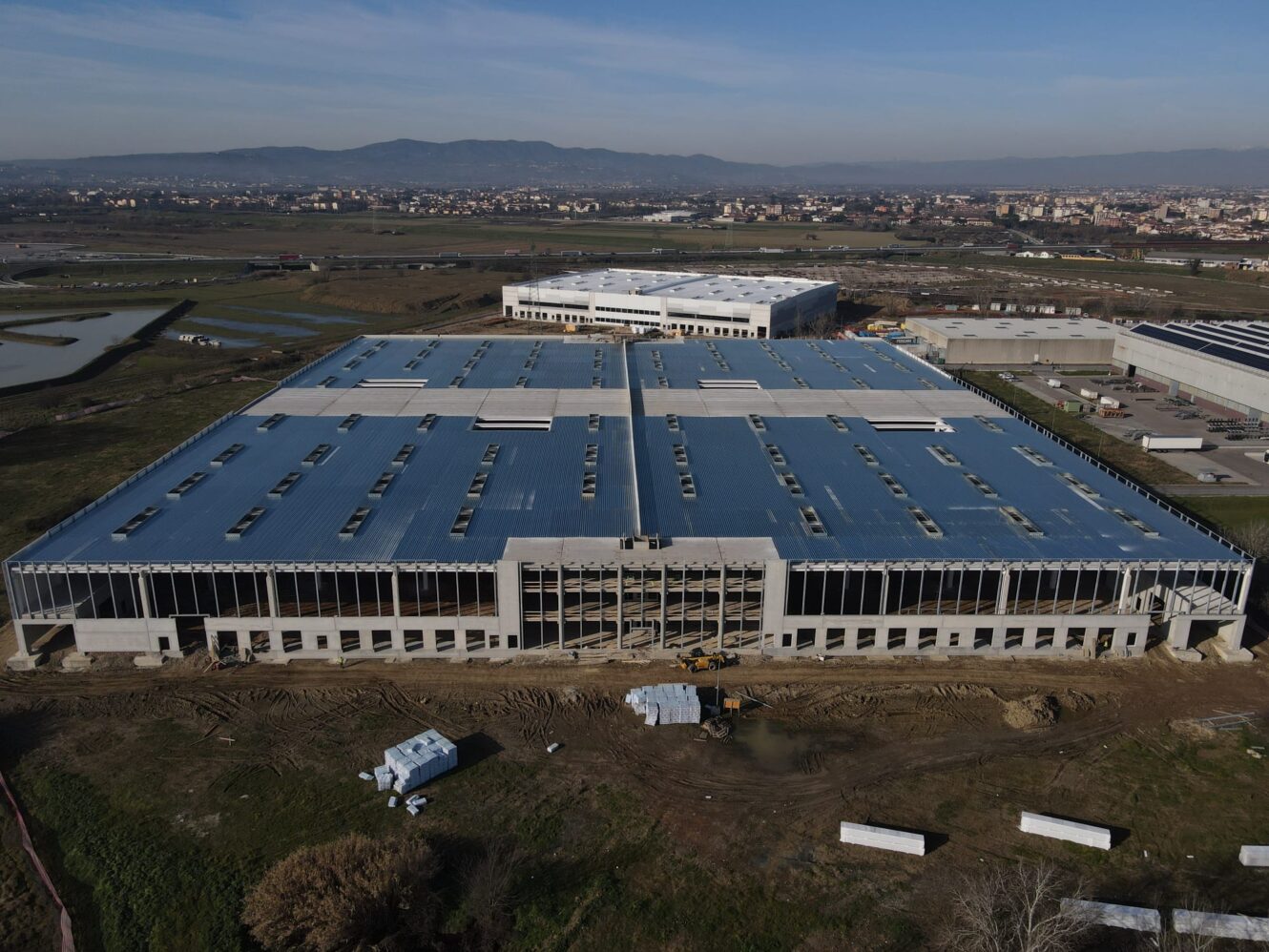 Veduta aerea di un grande capannone industriale parzialmente costruito con tetto metallico, circondato da terreni aperti e altri edifici industriali.