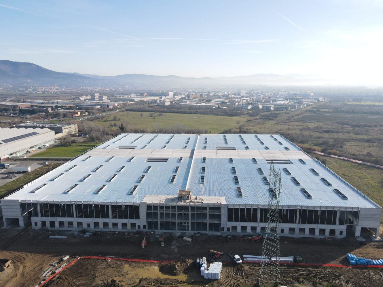 Veduta aerea di un grande capannone industriale in costruzione, con materiali da costruzione e attrezzature visibili a terra, sullo sfondo di campi e montagne lontane.