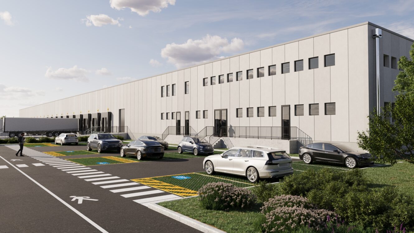 Un moderno magazzino industriale con diverse auto parcheggiate davanti, un semirimorchio su una banchina di carico e una persona che cammina vicino all'edificio sotto un cielo parzialmente nuvoloso.