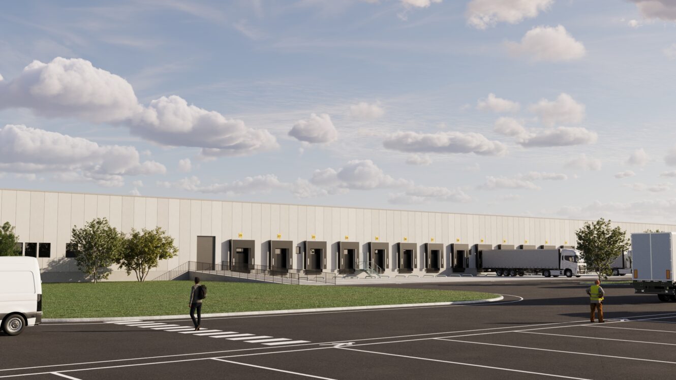 Un grande magazzino con banchine di carico, camion parcheggiati per il carico e lo scarico, un furgone bianco, due persone e un passaggio pedonale in primo piano sotto un cielo parzialmente nuvoloso.