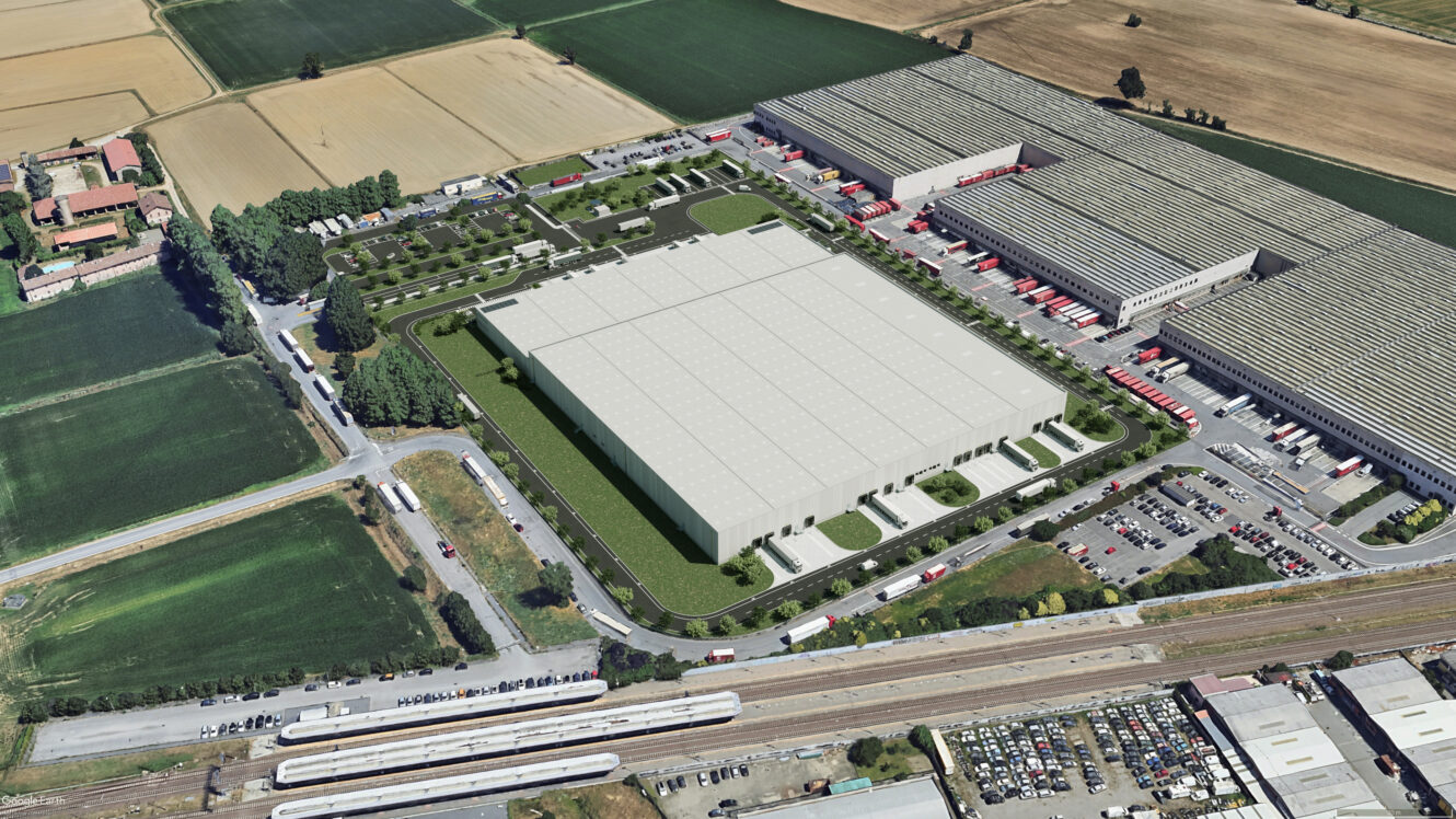 Veduta aerea di un grande complesso industriale con diversi edifici adibiti a magazzino, parcheggi, camion, strade circostanti e campi agricoli adiacenti.