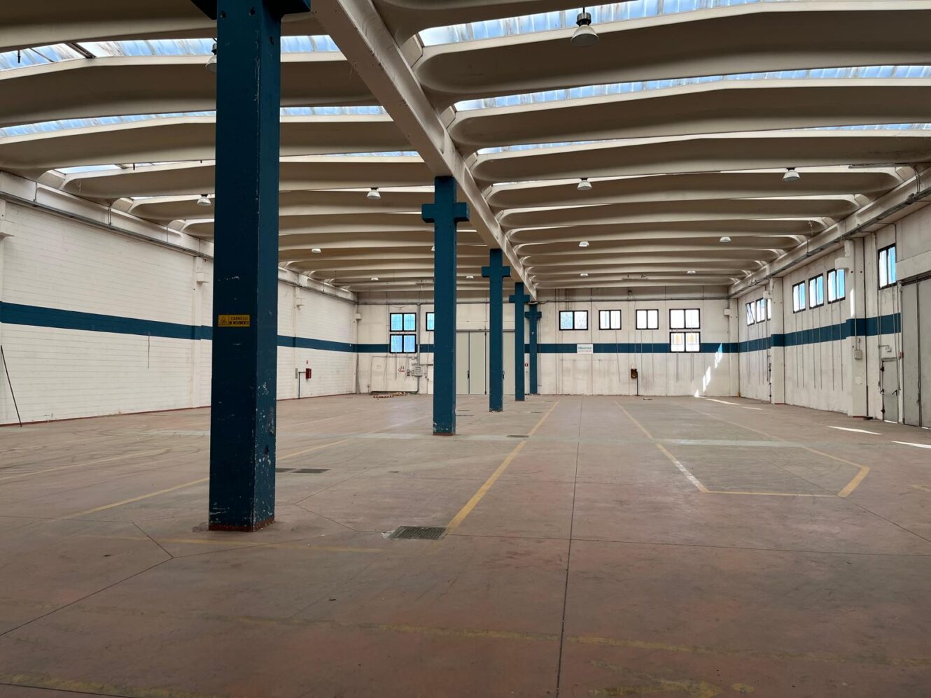 Interno di un ampio magazzino industriale vuoto, caratterizzato da soffitti alti, colonne di sostegno blu e luce naturale proveniente da lucernari.