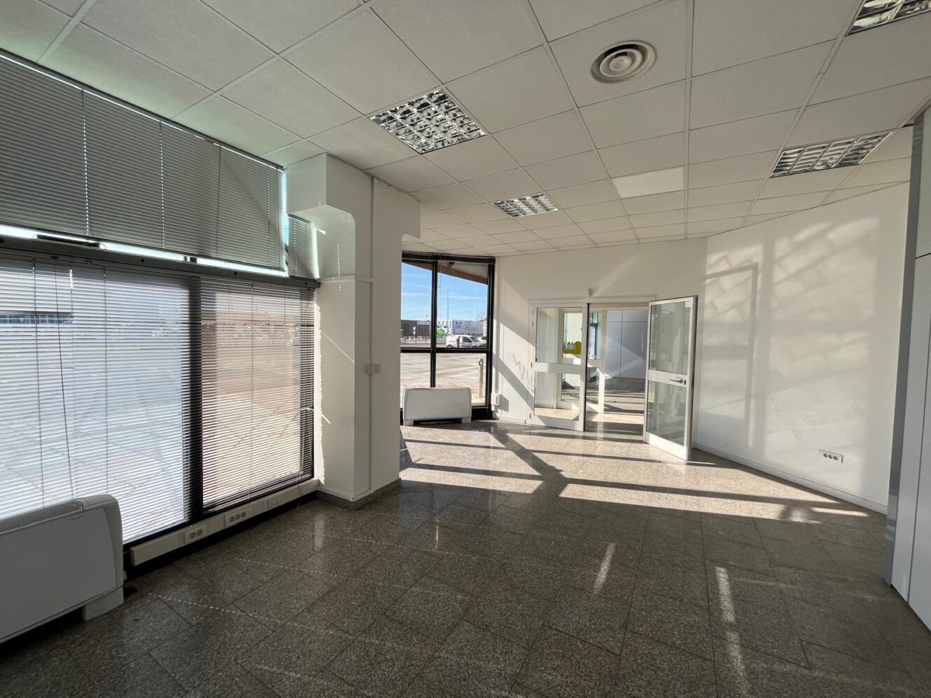 Ufficio vuoto con grandi finestre, porte di vetro, pavimento piastrellato e luci a soffitto; la luce del sole proietta ombre sulle pareti e sul pavimento.