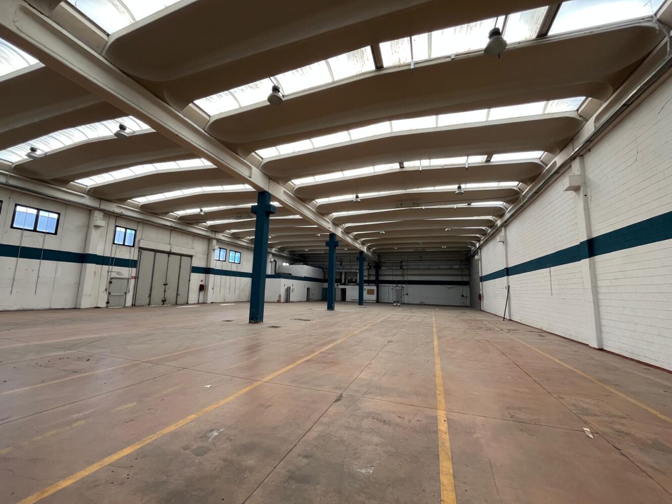 Ampio magazzino industriale vuoto con soffitti alti, travi a vista, lucernari e segni sul pavimento. Pareti bianche con una striscia blu.