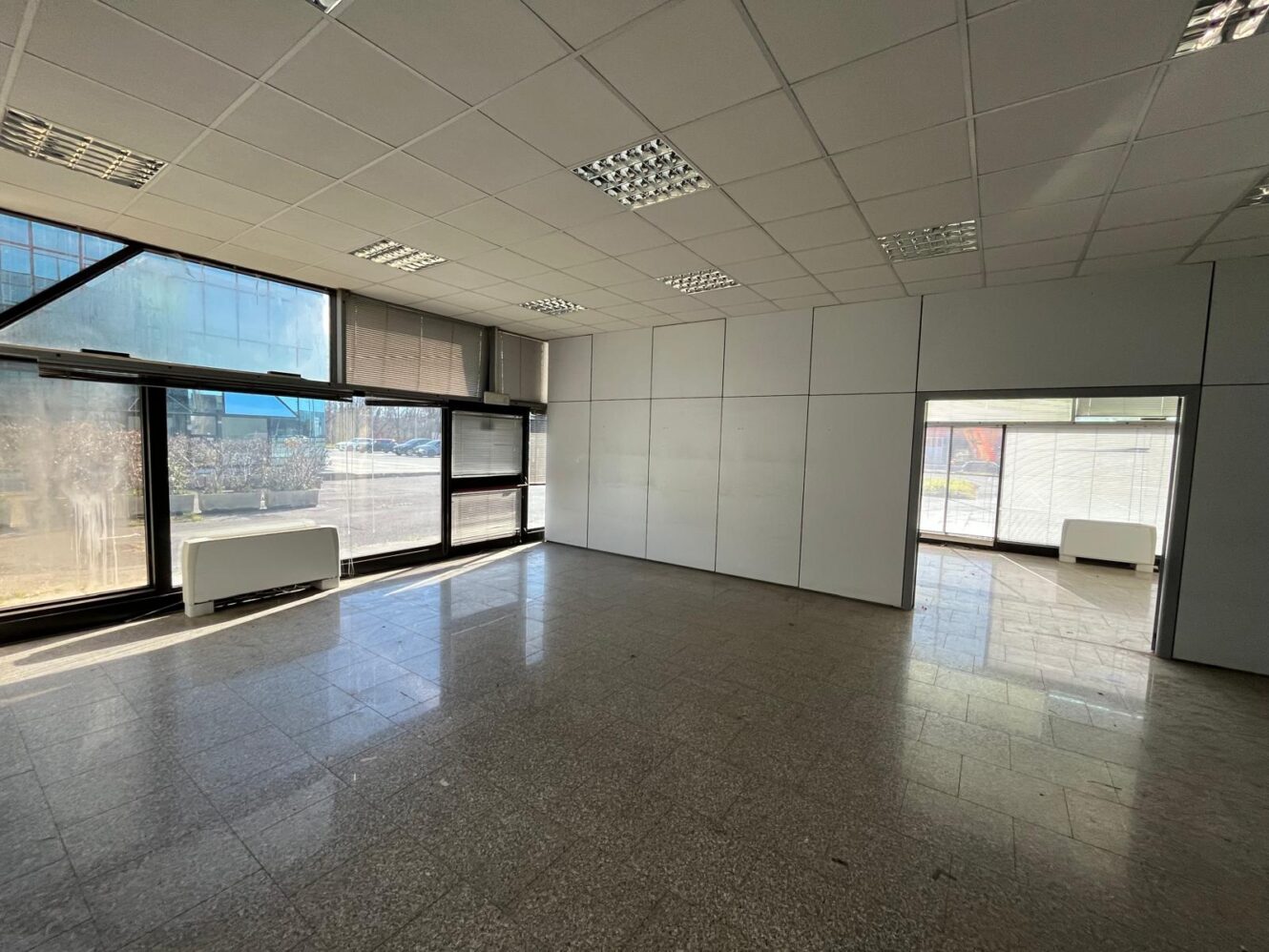 Ufficio vuoto con grandi finestre, pavimenti piastrellati, pareti bianche, luci fluorescenti e due porte aperte.