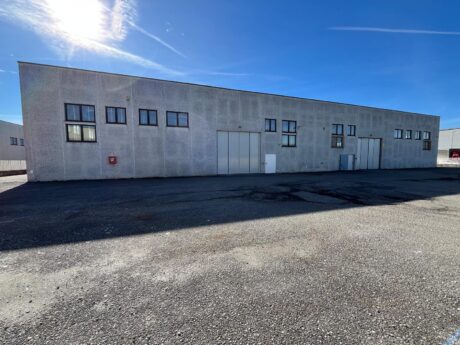 Un grande capannone industriale con molteplici finestre e porte si trova su un terreno asfaltato sotto un cielo azzurro e limpido.