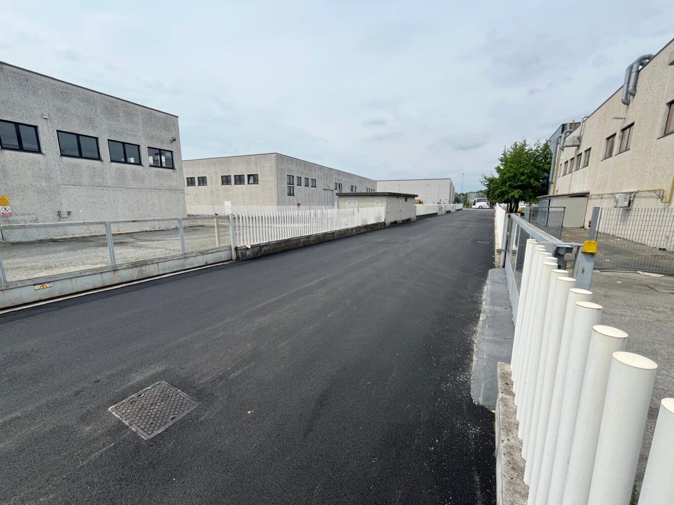 Una strada appena asfaltata corre tra edifici industriali con recinzioni bianche su ogni lato, sotto un cielo coperto.