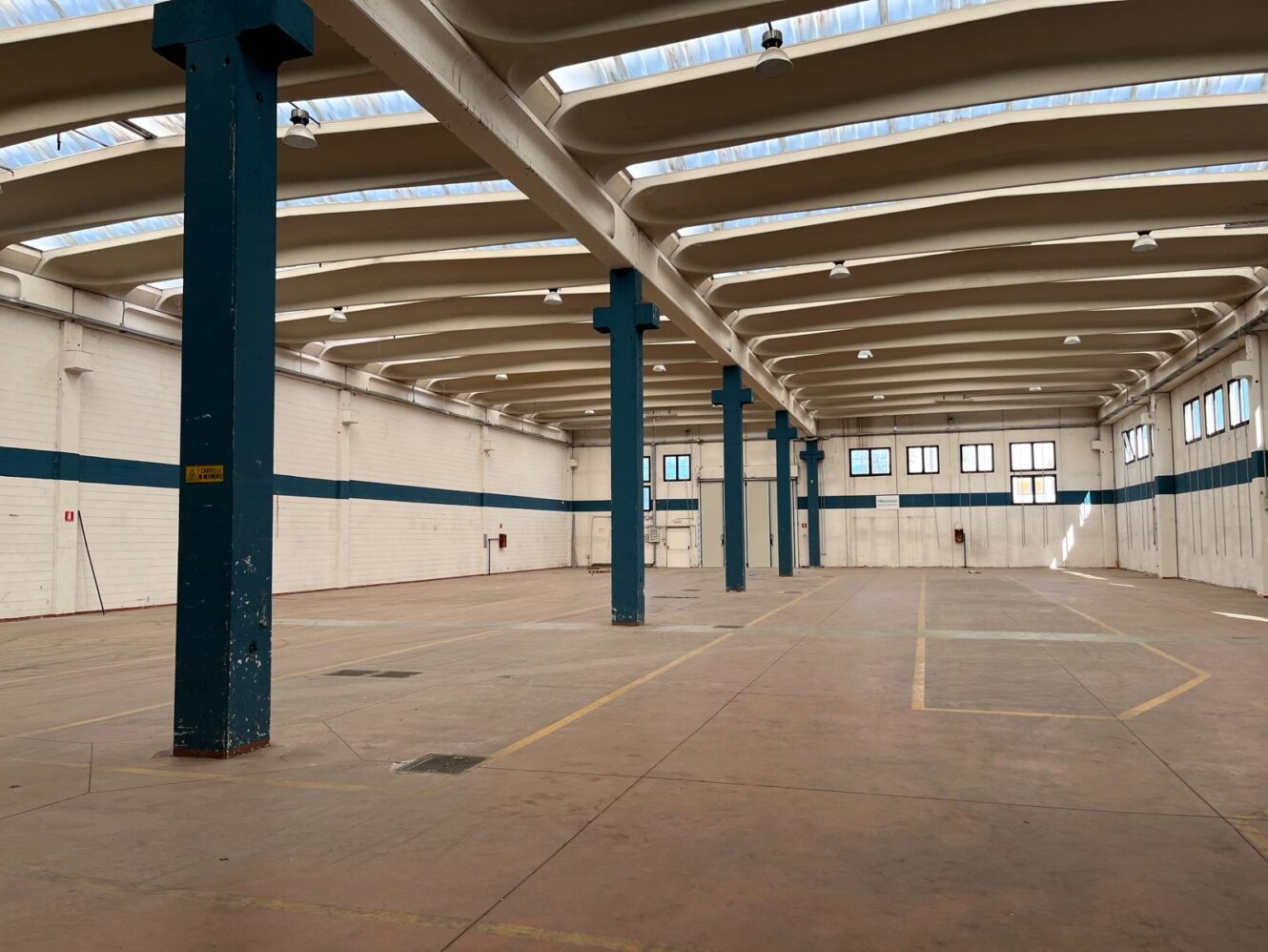 Ampio magazzino vuoto con soffitti alti, colonne portanti blu, pareti in mattoni bianchi, ampie finestre e pavimenti in cemento.