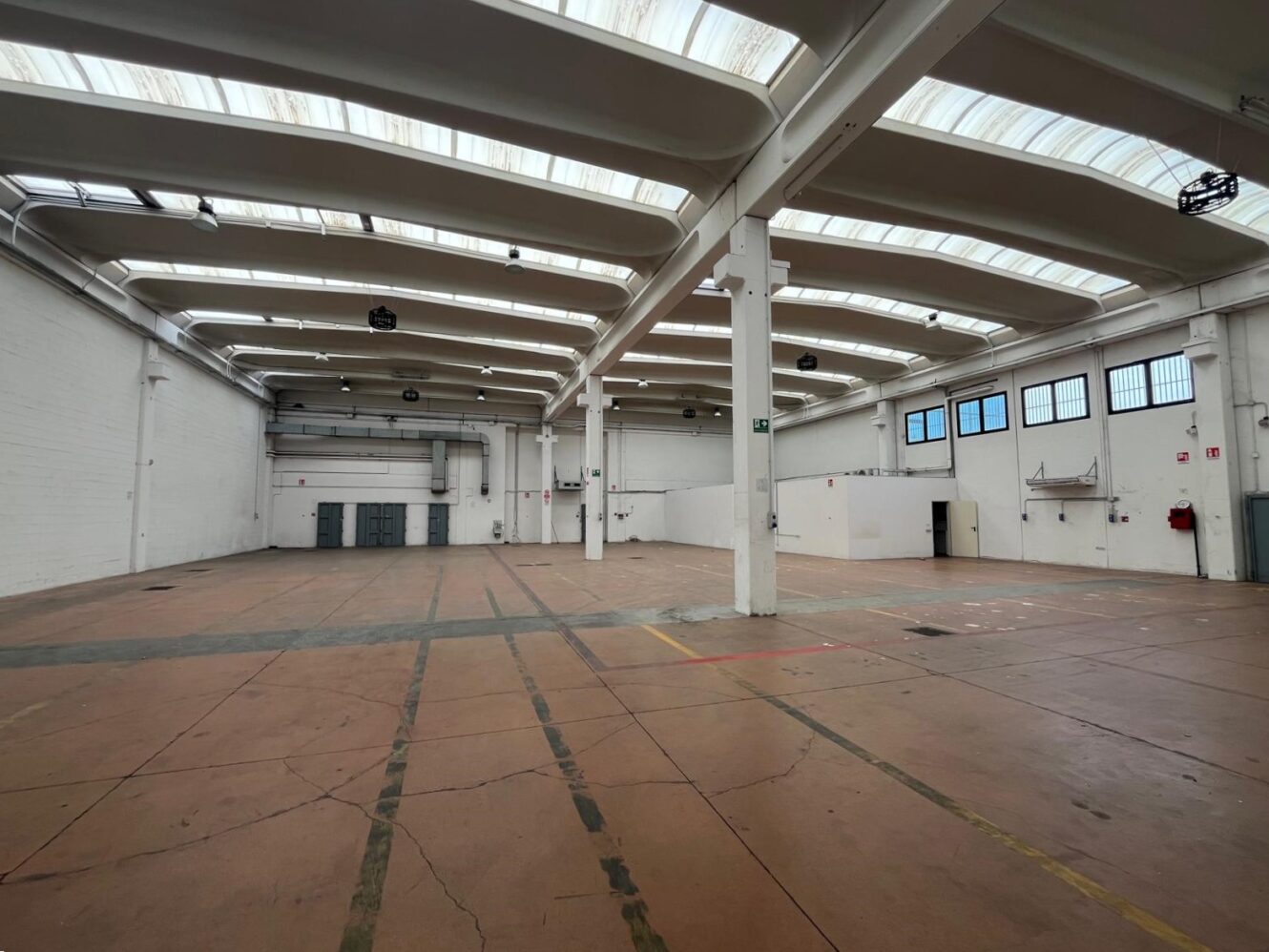 Magazzino industriale vuoto con soffitti alti, lucernari, pareti bianche e pavimenti in cemento. Porte e pannelli di servizio rivestono le pareti.