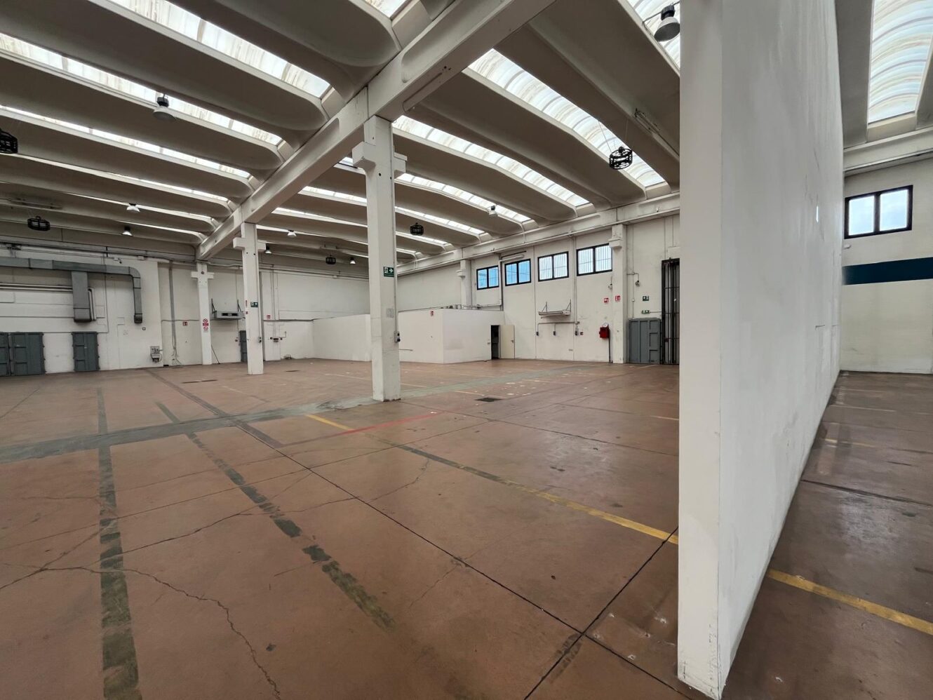 Ampio magazzino industriale vuoto con soffitti alti, travi a vista, lucernari, pavimenti in cemento, pareti bianche e finestre.
