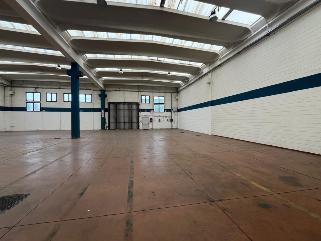 Un grande magazzino industriale vuoto con soffitti alti, lucernari, pareti bianche e un'ampia porta di garage in fondo.