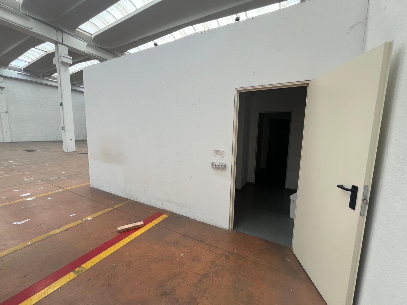 Una stanza bianca con una porta aperta all'interno di un grande magazzino con soffitti alti e lucernari; pavimento marrone con strisce rosse e gialle.