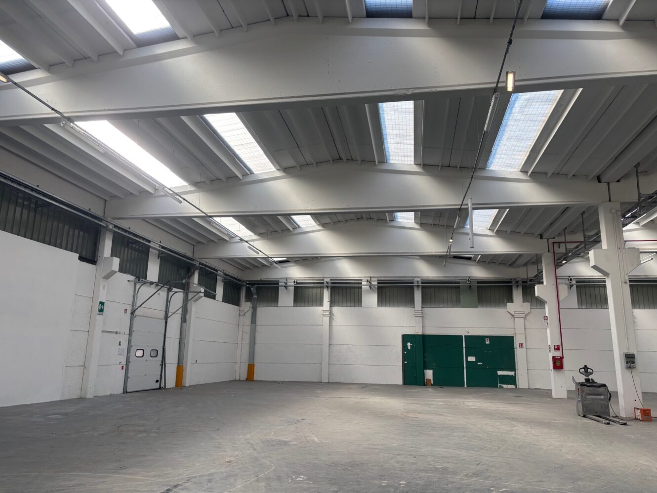 Magazzino industriale vuoto con soffitti alti, lucernari, pareti bianche, porte verdi e alcune attrezzature di servizio lungo la parete destra.