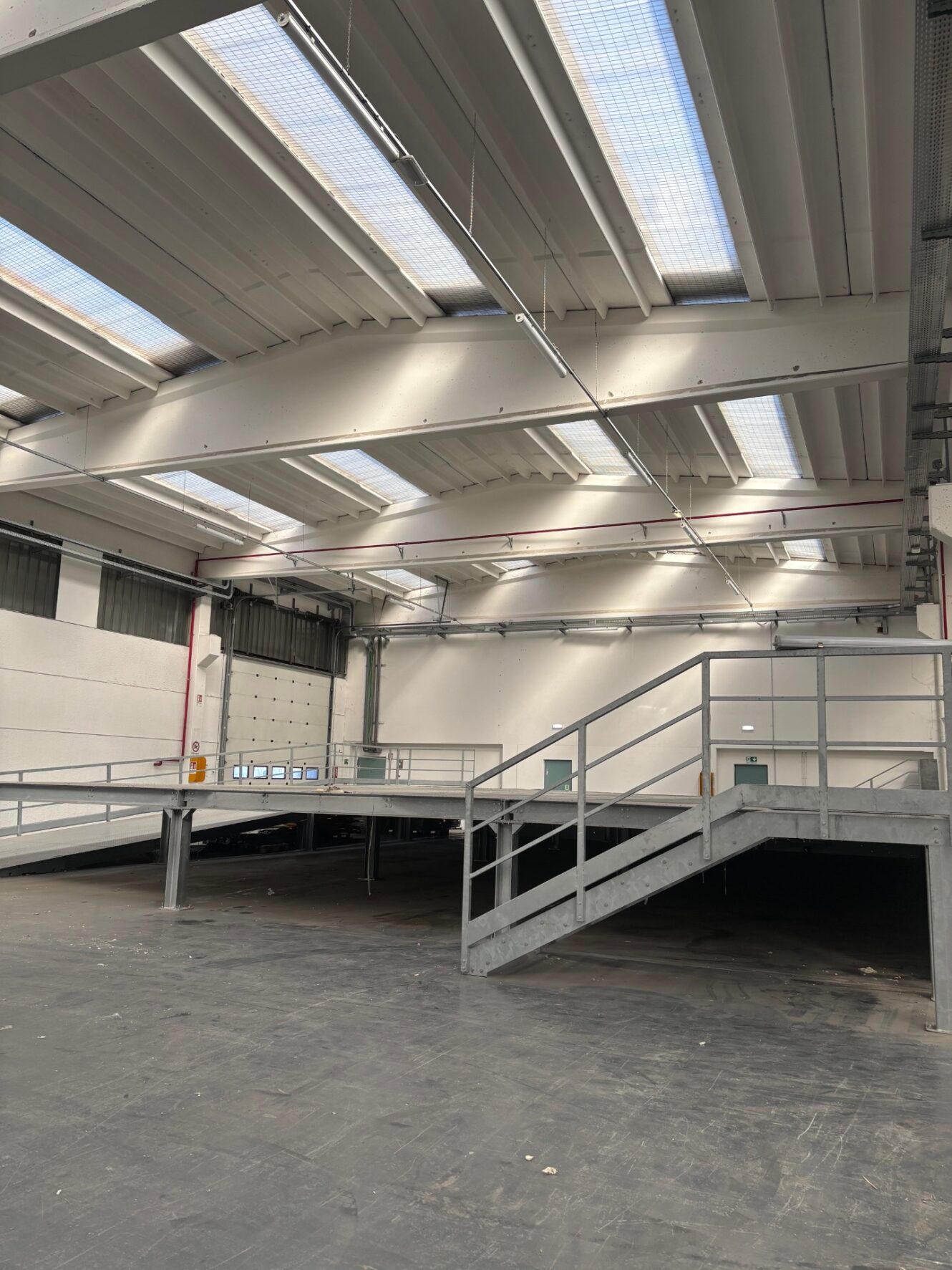 Grande magazzino industriale vuoto con piattaforma metallica soppalcata, scale, soffitti alti e lucernari. Le pareti sono bianche e il pavimento è in cemento scuro.