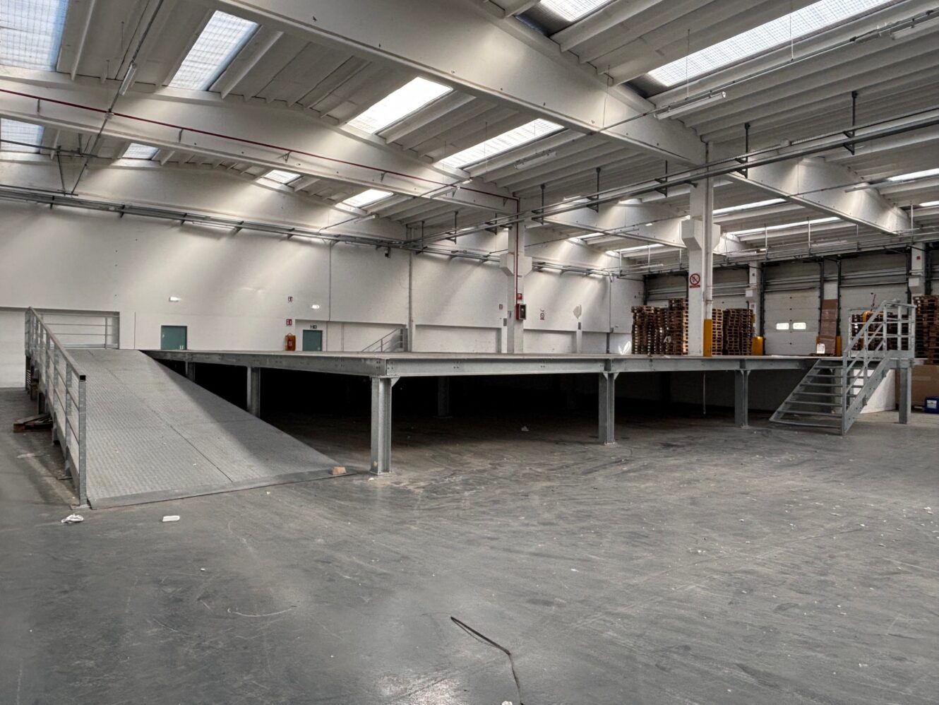 Ampio magazzino industriale vuoto con piattaforma metallica soppalcata, rampe di accesso e scale sotto un soffitto alto con lucernari.